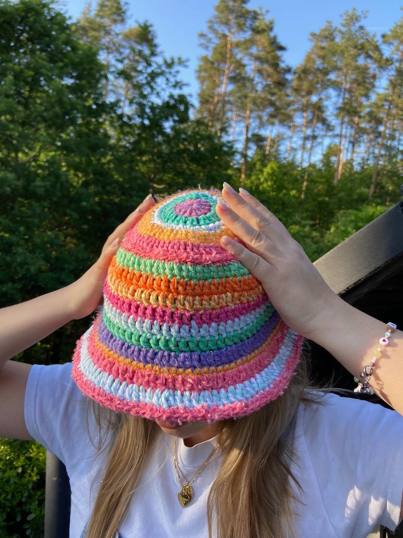 May include: A colorful crocheted bucket hat with stripes of pink, orange, green, purple, and white. The hat has a round, flat crown and a wide brim.