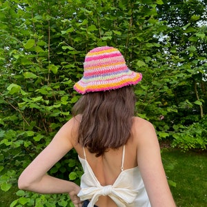 May include: A person wearing a white tank top with a bow tie in the back and a colorful crocheted bucket hat.