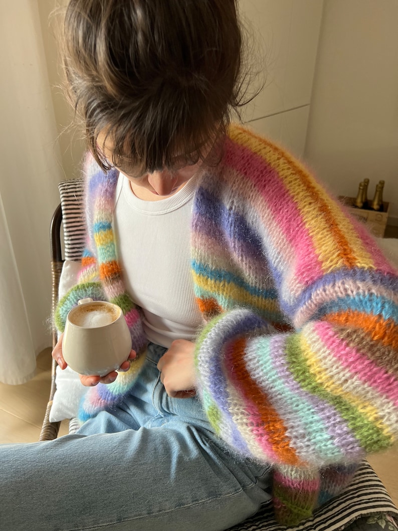 May include: A person wearing a rainbow striped fuzzy cardigan with a white vest top and blue jeans. They are holding a white ceramic mug with a light brown drink.