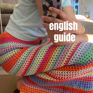 May include: A person wearing a light blue shirt and a colorful crochet skirt. The skirt is made of a variety of colors, including pink, orange, yellow, green, and blue. The skirt is crocheted in a pattern of stripes and squares. The person is holding a cell phone with a floral case. The text "english guide" is written on the image.