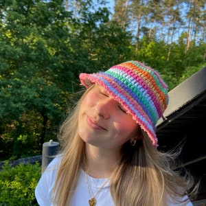 May include: A person wearing a rainbow striped crochet bucket hat. The hat has a pink brim and is made with a variety of colorful yarns.