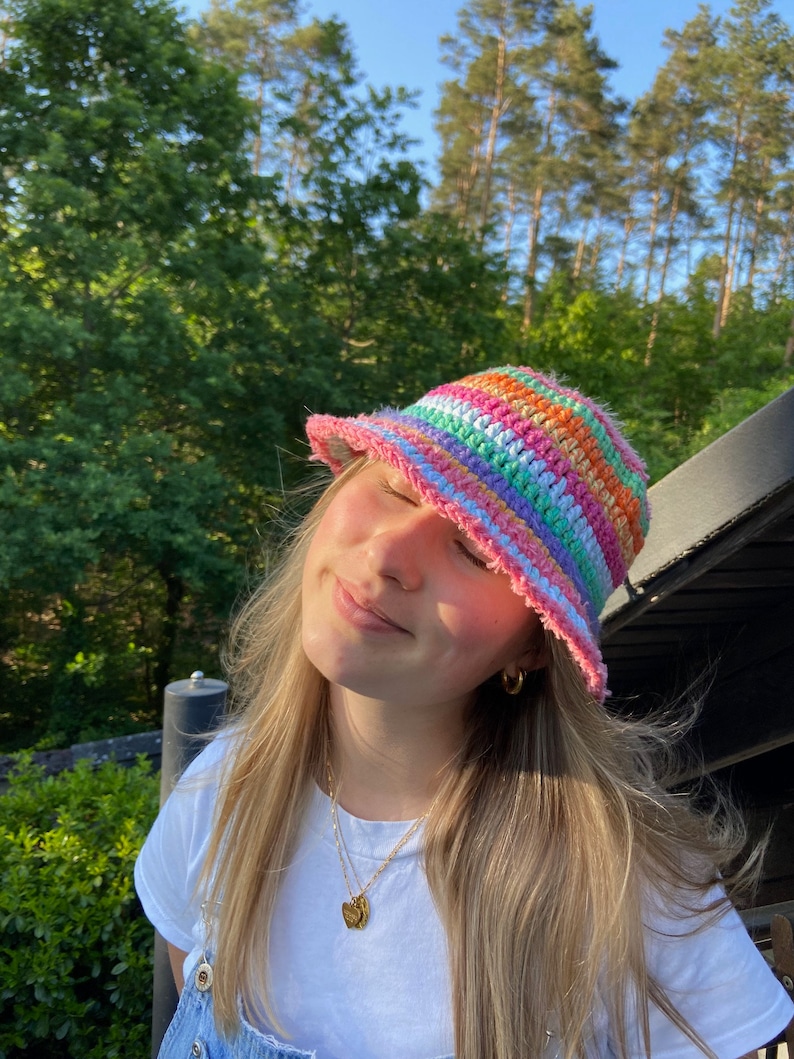 May include: A person wearing a rainbow striped crochet bucket hat. The hat has a pink brim and is made with a variety of colorful yarns.
