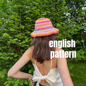 May include: A person wearing a colorful crocheted bucket hat with pink, yellow, and green stripes. The hat has a wide brim and is made with a textured yarn. The person is wearing a white tank top with a bow tied in the back and black pants. The text "english pattern" is written in white letters on a black background.