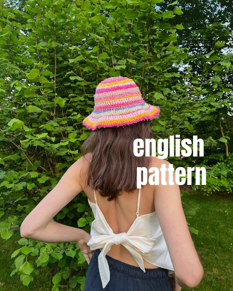 May include: A person wearing a colorful crocheted bucket hat with pink, yellow, and green stripes. The hat has a wide brim and is made with a textured yarn. The person is wearing a white tank top with a bow tied in the back and black pants. The text "english pattern" is written in white letters on a black background.