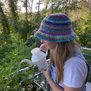 May include: A person wearing a colorful crocheted bucket hat waters plants with a white watering can.