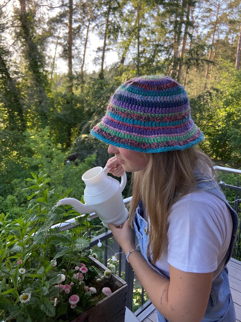 May include: A person wearing a colorful crocheted bucket hat waters plants with a white watering can.