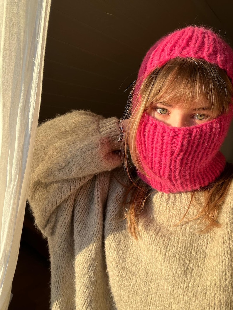 May include: A woman wearing a pink knitted hat and matching neck warmer. The hat and neck warmer are both made of a soft, fluffy yarn. The woman is wearing a beige sweater.