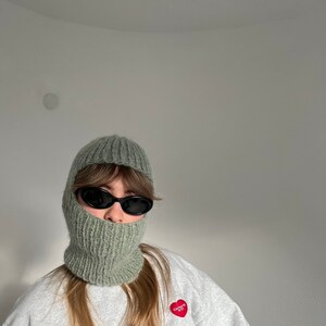 May include: A person wearing a green knitted balaclava with black sunglasses and a grey sweatshirt with a red heart patch that says "Calvert W.P."