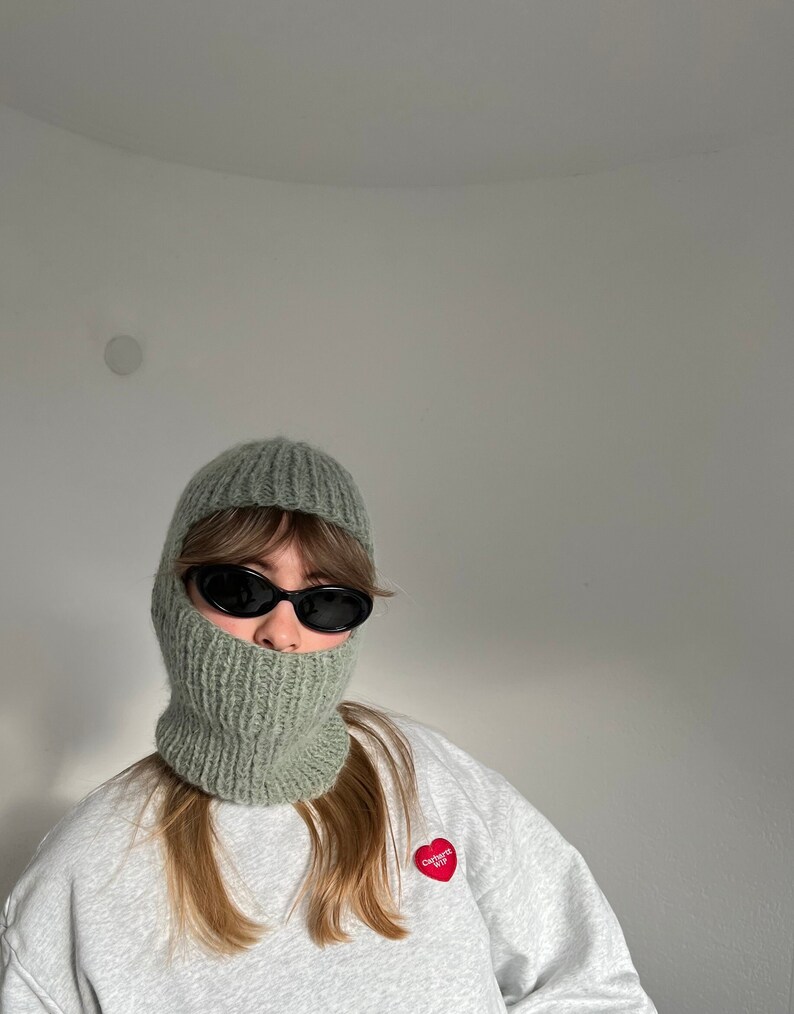 May include: A person wearing a green knitted balaclava with black sunglasses and a grey sweatshirt with a red heart patch that says "Calvert W.P."