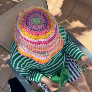 May include: A person wearing a colorful crocheted bucket hat and a green and white striped shirt is sitting in a chair and knitting with green yarn.