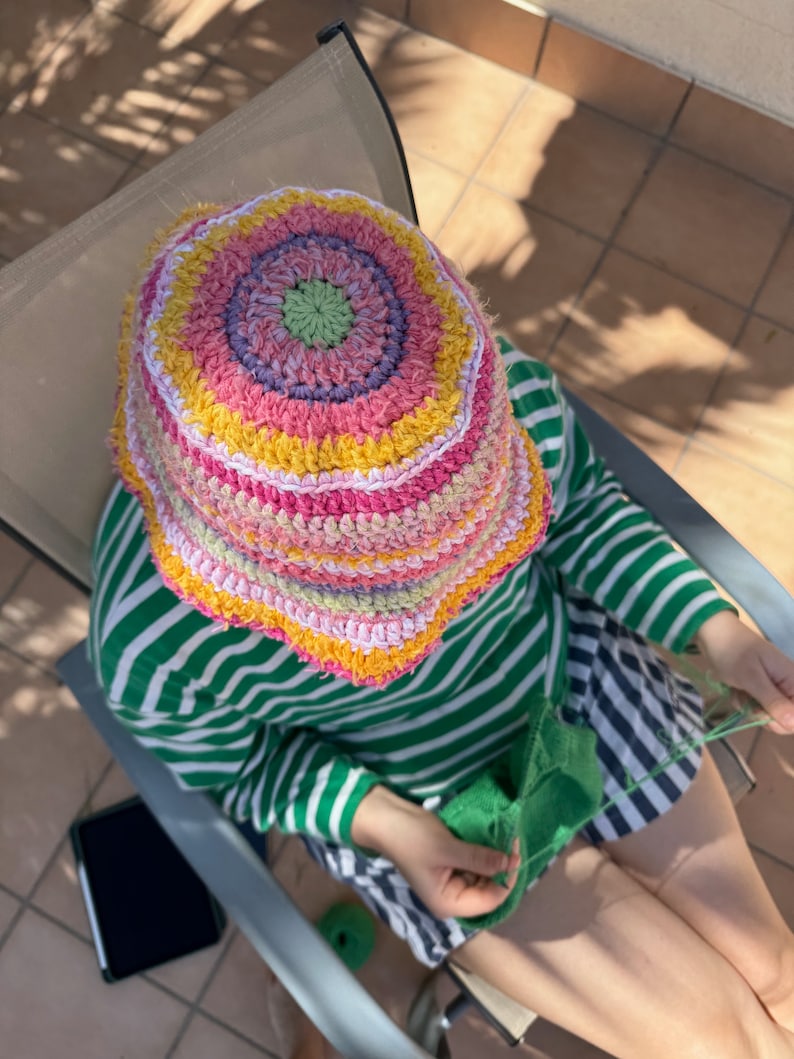 May include: A person wearing a colorful crocheted bucket hat and a green and white striped shirt is sitting in a chair and knitting with green yarn.