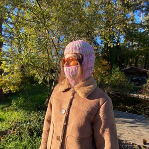 May include: A person wearing a pink and white knitted balaclava with a brown faux fur lined jacket. The person is standing in a wooded area with a pond in the background.