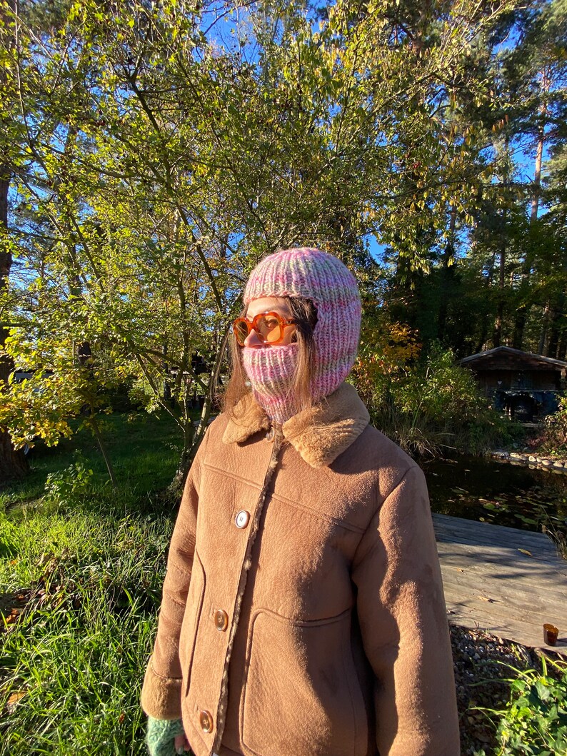 May include: A person wearing a pink and white knitted balaclava with a brown faux fur lined jacket. The person is standing in a wooded area with a pond in the background.