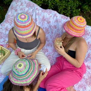 May include: Three friends are sitting on a blanket outdoors, each wearing a colorful crocheted bucket hat. They are working on crochet projects, with yarn and crochet hooks visible.
