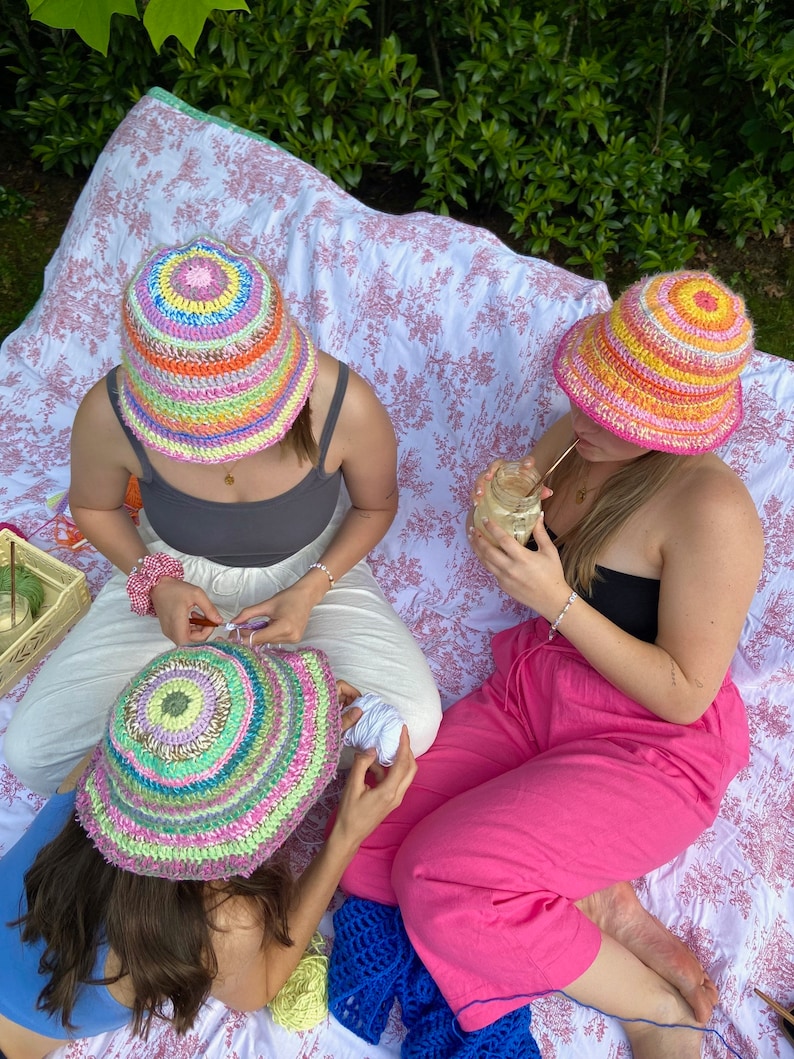 May include: Three friends are sitting on a blanket outdoors, each wearing a colorful crocheted bucket hat. They are working on crochet projects, with yarn and crochet hooks visible.