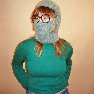 May include: A person wearing a green long-sleeved shirt, blue jeans, and a light green knitted balaclava.