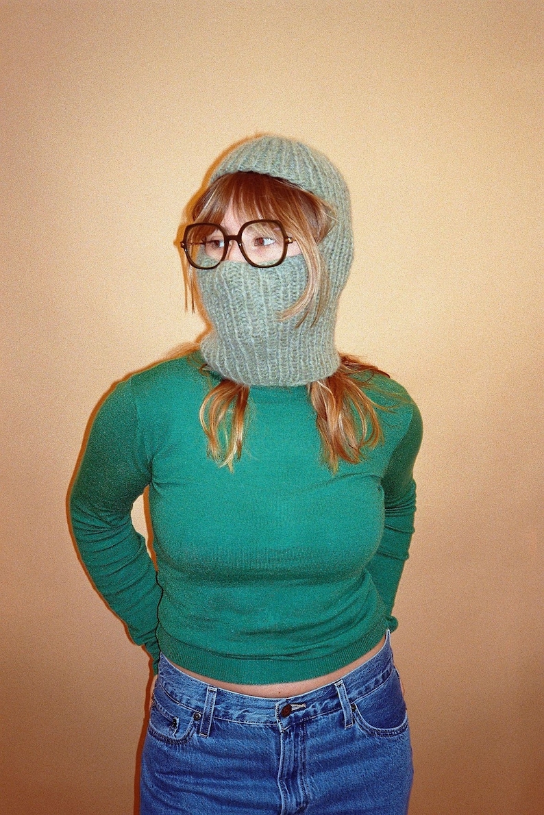 May include: A person wearing a green long-sleeved shirt, blue jeans, and a light green knitted balaclava.