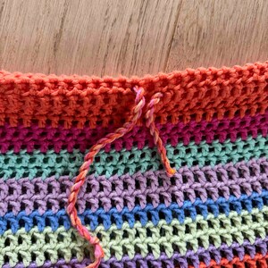 K&ouml;nnte beinhalten: Nahaufnahme eines mehrfarbigen H&auml;kelstoffs mit einem gestreiften Muster in Rosa, Orange und Gr&uuml;n. Der Stoff wird mit einem langen, d&uuml;nnen, orangenen Faden zusammengen&auml;ht.