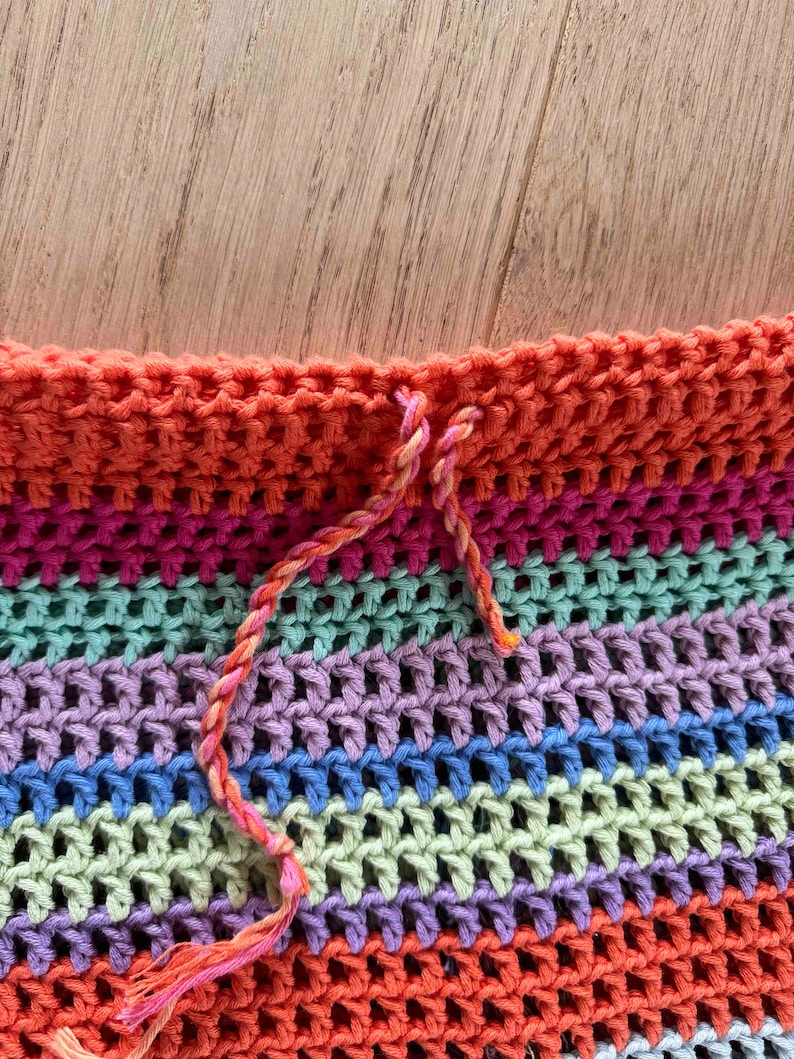 K&ouml;nnte beinhalten: Nahaufnahme eines mehrfarbigen H&auml;kelstoffs mit einem gestreiften Muster in Rosa, Orange und Gr&uuml;n. Der Stoff wird mit einem langen, d&uuml;nnen, orangenen Faden zusammengen&auml;ht.