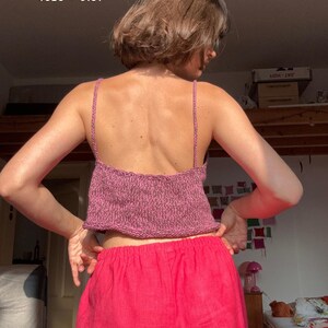 May include: A knitted pink crop top with thin straps is worn with bright pink pants. The text "SANDNES GARN LINE 4626 + 3161" is visible at the top of the image. The person is facing away from the camera.