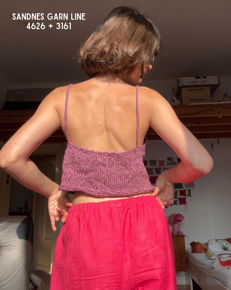 May include: A knitted pink crop top with thin straps is worn with bright pink pants. The text "SANDNES GARN LINE 4626 + 3161" is visible at the top of the image. The person is facing away from the camera.