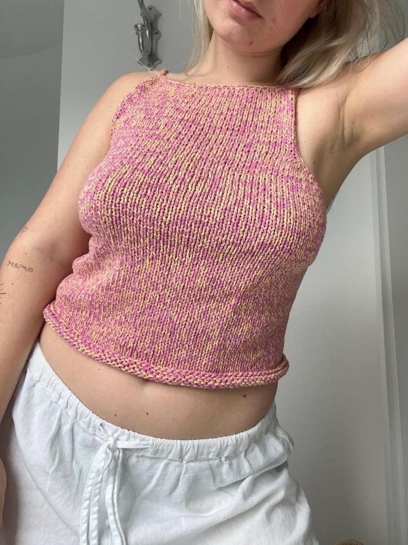 May include: A pink and yellow knitted tank top. The top has a square neckline and thin straps. The knitted fabric has a textured appearance. The bottom hem is a solid pink color. The tank top is a summer garment.