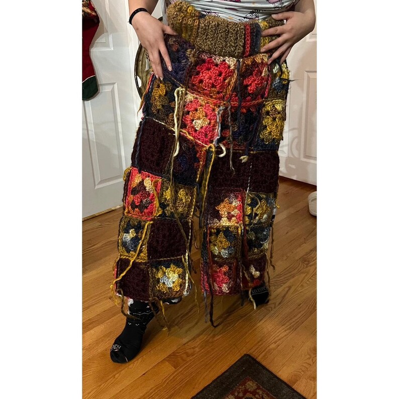 May include: Handmade crochet pants in a patchwork design. The pants feature a variety of colors including red, yellow, blue, and brown. The squares are joined together with visible yarn ends. The waist band is a solid brown color.