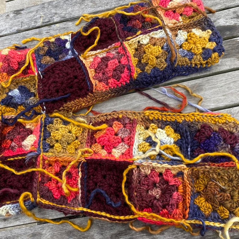 May include: Close-up of two unfinished crochet pieces. The pieces are made of colorful granny squares in shades of red, yellow, blue, and brown. Loose yarn strands in yellow and brown are visible, indicating the project is in progress. The pieces are resting on a wooden surface.