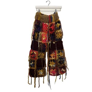 May include: Handmade crochet pants in a patchwork design. The pants feature a variety of colors, including red, yellow, blue, and brown. The pants are hanging on a white hanger. The pants have a high waist and a wide leg.