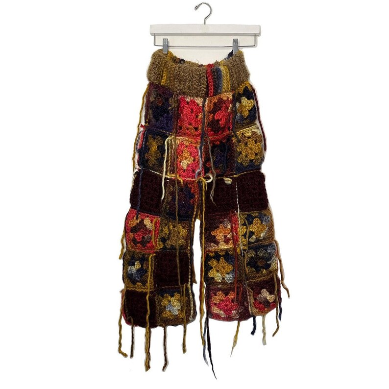 May include: Handmade crochet pants in a patchwork design. The pants feature a variety of colors, including red, yellow, blue, and brown. The pants are hanging on a white hanger. The pants have a high waist and a wide leg.