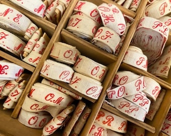 May include: A wooden box filled with rolls of off-white ribbon. The ribbon is printed with red lettering, including initials and numbers. The rolls are neatly organized within the box's compartments, showcasing a vintage aesthetic.
