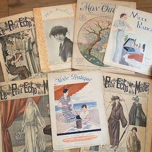 May include: A collection of vintage fashion magazines with colorful covers. The magazines feature illustrations of women in various outfits, with titles like "Mode Illustrée" and "Le Petit Echo de la Mode". The magazines are arranged on a wooden surface.