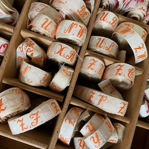 Könnte beinhalten: Eine Holzkiste gefüllt mit Rollen aus cremefarbenem Band. Das Band ist mit orangefarbenen und roten Buchstaben und Initialen bestickt. Die Kiste hat mehrere Fächer, und die Bänder sind ordentlich angeordnet.