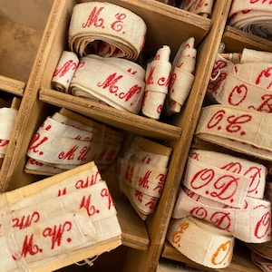Może przedstawiać: Zbliżenie drewnianej skrzynki wypełnionej rolkami kremowej wstążki. Wstążka jest zadrukowana czerwonymi literami, z inicjałami i nazwiskami. Skrzynka ma wiele przegródek, a wstążki są starannie ułożone.