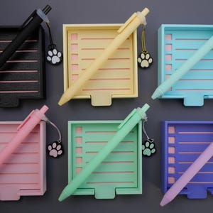 May include: A collection of colorful pens and matching notepads. The pens are black, yellow, teal, pink, green, and purple. Each pen rests on a notepad of the same color, with a paw print charm attached. The notepads have a lined design.