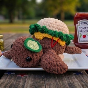 May include: A crocheted turtle plush with a brown shell, a tan bun top, green lettuce, and yellow cheese layers, resembling a hamburger. The turtle has a small green circle on its head and black eyes, resting on a white square plate next to a bottle of Heinz Tomato Ketchup.
