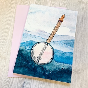 May include: A watercolor painting of a banjo with a blue and white background. The banjo is drawn in black ink and has a light pink interior. The background is a watercolor wash of blue and white.