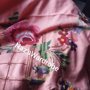 May include: A pink textile with cross-stitched floral embroidery. The design features a large red rose, green leaves, and various other colorful flowers. The text "Naz's Wardrobe" is visible on the textile.
