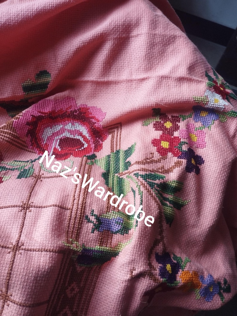 May include: A pink textile with cross-stitched floral embroidery. The design features a large red rose, green leaves, and various other colorful flowers. The text "Naz's Wardrobe" is visible on the textile.