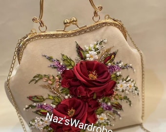 May include: A white velvet handbag with a gold chain strap and a gold clasp. The bag is decorated with a floral design of red roses, white flowers, and green leaves. The text "Naz's Wardrobe" is embroidered on the bag.