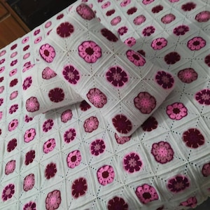 Op de afbeelding: Een wit gehaakt deken met roze en bordeauxrode bloemen. Het deken wordt geleverd met een bijpassend kussen.