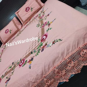 May include: A light pink bedspread and two pillowcases with floral embroidery. The bedspread features a detailed floral design with a crocheted edge. The pillowcases have a rose design. The text "Naz'sWardrobe" is visible on the bedspread.