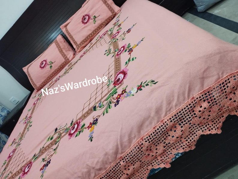 May include: A light pink bedspread and two pillowcases with floral embroidery. The bedspread features a detailed floral design with a crocheted edge. The pillowcases have a rose design. The text "Naz'sWardrobe" is visible on the bedspread.