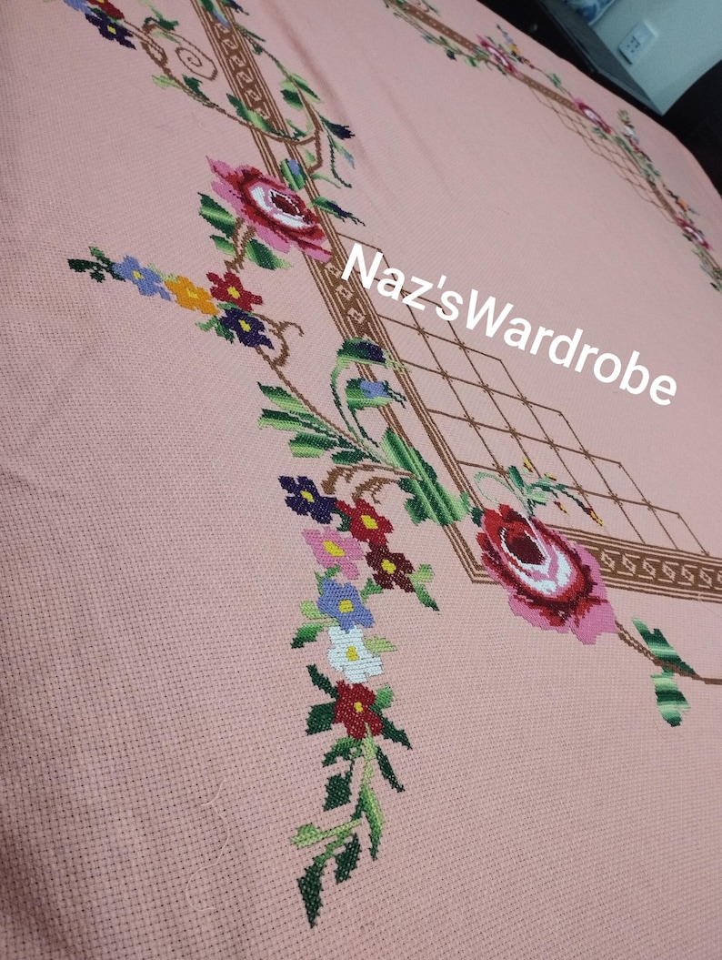May include: A pink tablecloth with embroidered floral designs. The embroidery features red, pink, and white roses, along with other colorful flowers and green leaves. The text "Naz's Wardrobe" is visible.