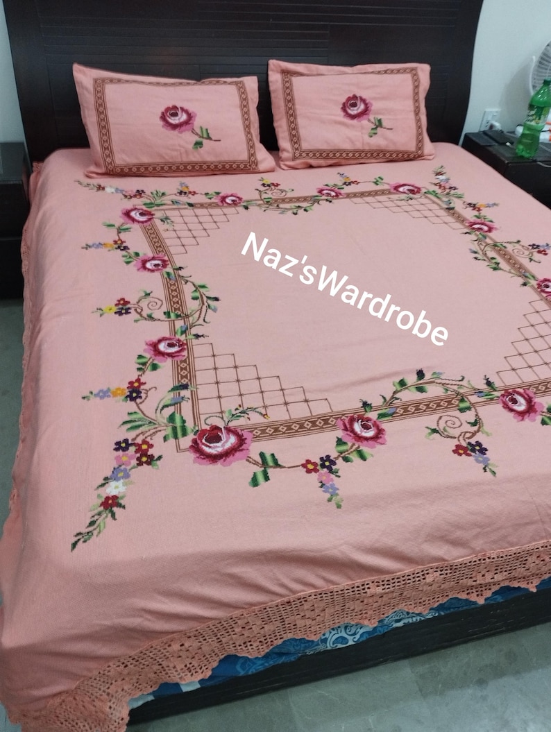 May include: A pink bedspread and two pillowcases with embroidered floral designs. The bedspread features a large square border with a floral pattern and the text "Naz'sWardrobe". The pillowcases have a single rose design.