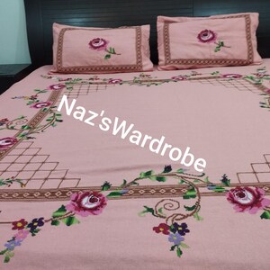May include: Pink embroidered bedspread and two pillowcases with a floral design. The bedspread features a large central floral pattern with a border of geometric shapes and floral vines. The pillowcases have a single rose design. The text "Naz's Wardrobe" is visible.