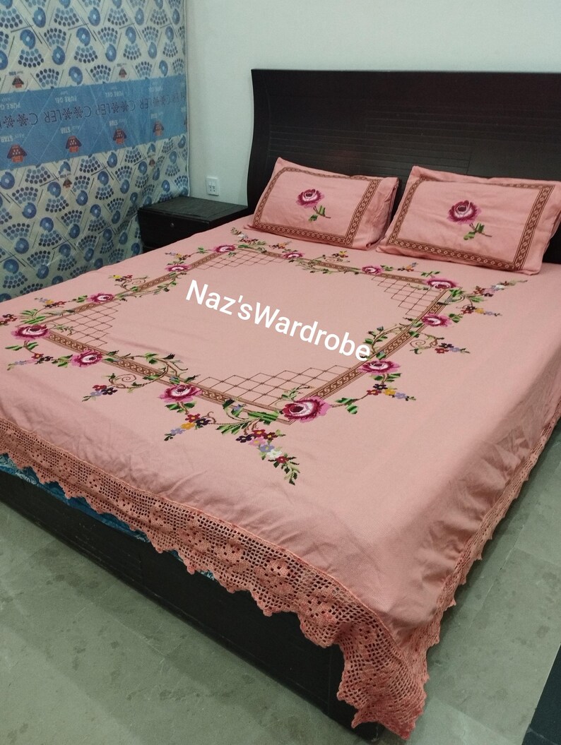 May include: A pink bedspread and two pillowcases with floral embroidery. The bedspread features a large embroidered square design with roses and other flowers. The pillowcases have a single rose embroidered in the center. The bedspread has a crocheted lace edge. The text "Naz's Wardrobe" is visible.
