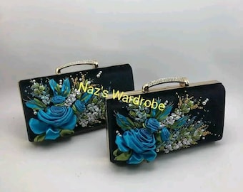 May include: Two black satin evening clutches with gold metal frames and handles. Each clutch features a floral design with blue and silver ribbon embroidery. The text "Naz's Wardrobe" is visible on the left clutch.