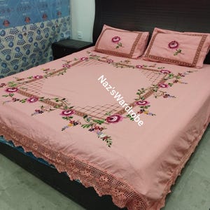 May include: A pink bedspread and two pillowcases with embroidered floral designs. The bedspread features a large square floral pattern and a scalloped lace edge. The text "Naz's Wardrobe" is visible on the bedspread.
