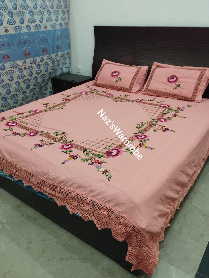 May include: A pink bedspread and two pillowcases with embroidered floral designs. The bedspread features a large square floral pattern and a scalloped lace edge. The text "Naz's Wardrobe" is visible on the bedspread.